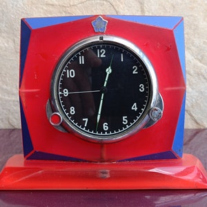 Può includere: Un orologio rosso e blu con un quadrante nero e numeri bianchi. L'orologio ha un bordo argentato e un piccolo pulsante rosso sul lato. L'orologio è posto su una base rossa.