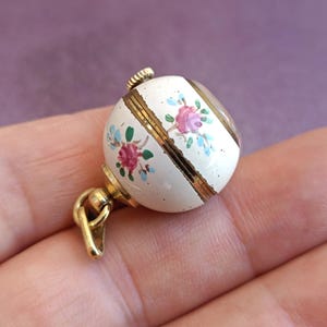 May include: A small, round, white pendant with gold accents and a floral design. The pendant features hand-painted pink and blue flowers. The pendant is attached to a gold-colored bail and clasp.