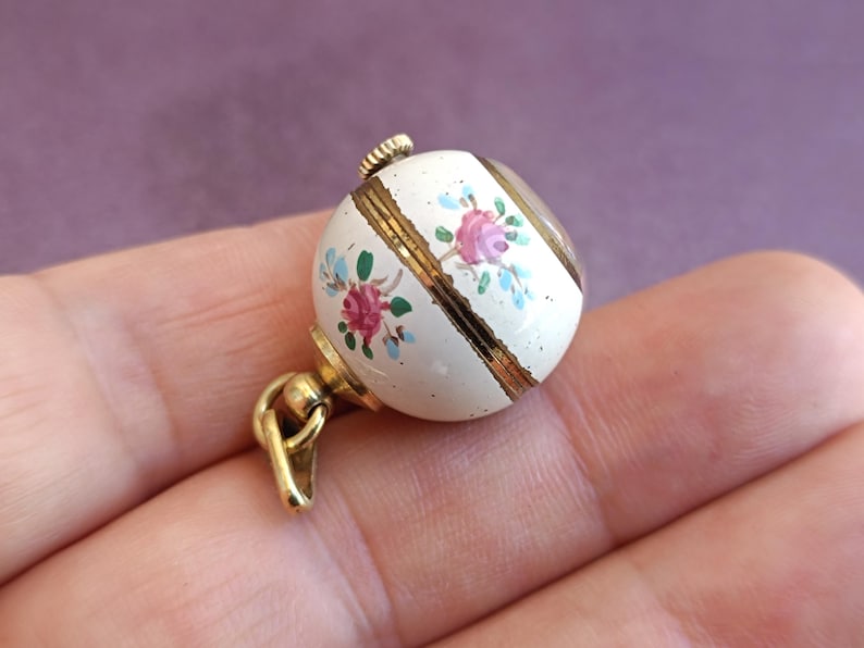 May include: A small, round, white pendant with gold accents and a floral design. The pendant features hand-painted pink and blue flowers. The pendant is attached to a gold-colored bail and clasp.