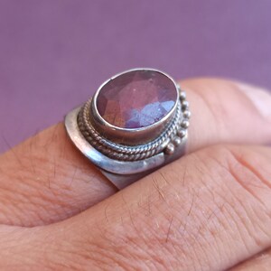 Pode incluir: Um anel de prata com uma grande pedra preciosa ovalada de cor p&uacute;rpura engastada em uma moldura de prata. O anel tem uma banda decorativa com uma borda de contas.