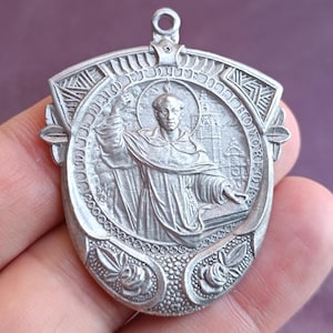 May include: Silver pendant with a detailed relief of a religious figure. The pendant features an oval central image surrounded by an ornate border with floral and geometric designs. The figure is depicted with a halo and is holding a book.