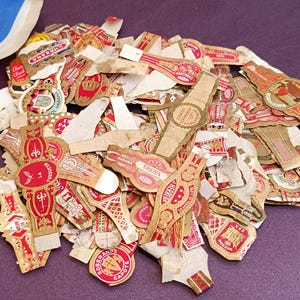 May include: A pile of assorted vintage cigar bands, predominantly in red, gold, and cream hues. The bands showcase detailed designs, text, and illustrations, indicating a historical or collectible nature. Some bands are torn or fragmented.