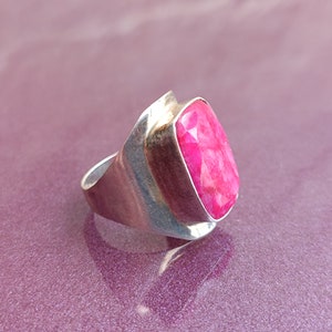 May include: A silver ring with a large, faceted, oval-shaped, bright pink gemstone. The ring has a wide, curved band and a bezel setting that holds the stone securely. The gemstone's vibrant colour contrasts with the silver.