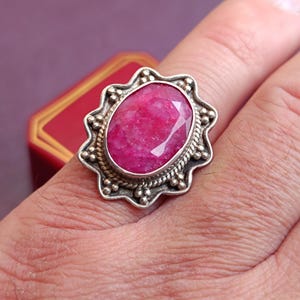 Op de afbeelding: Een zilveren ring met een grote, ovaal geslepen robijn. De robijn heeft een levendige roze kleur en is gezet in een decoratieve zilveren zetting met een geschulpte rand. De ring zit om een vinger.