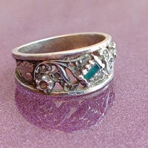 May include: A silver-toned metal ring featuring an intricate openwork design with floral motifs and a single rectangular green gemstone set horizontally. The band has a polished finish and a wide profile with detailed cutouts along the entire circumference.