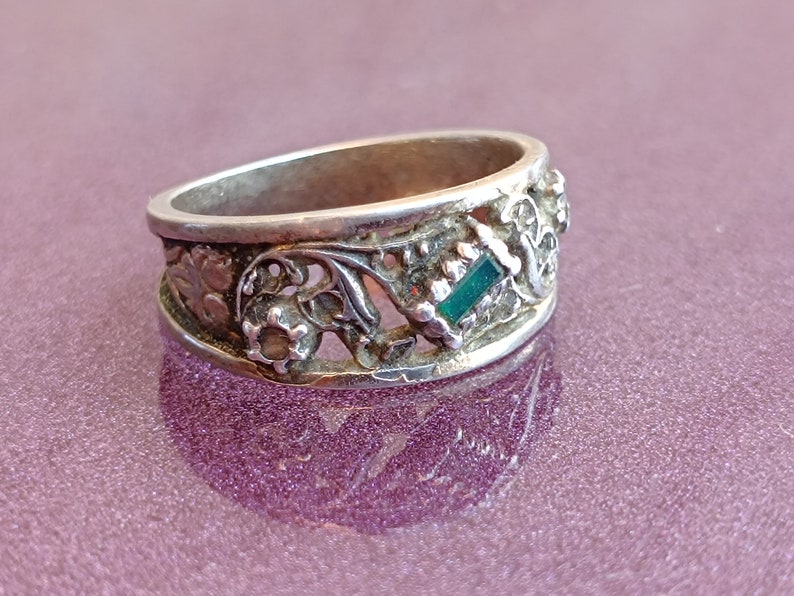 May include: A silver-toned metal ring featuring an intricate openwork design with floral motifs and a single rectangular green gemstone set horizontally. The band has a polished finish and a wide profile with detailed cutouts along the entire circumference.