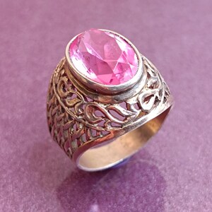 May include: A silver ring featuring a large, oval-cut pink gemstone. The ring's band has an intricate, open filigree pattern. The pink stone is set in a silver bezel. The ring is shown on a purple background.