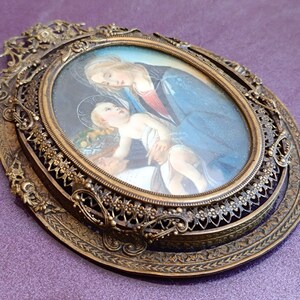 K&ouml;nnte beinhalten: Ein antikes ovales religi&ouml;ses Portr&auml;t in einem dekorativen Bronzerahmen. Das Kunstwerk zeigt eine Frau in einem blauen Gewand und ein Kind mit Heiligenschein. Der Rahmen hat komplizierte Filigran- und Blumendetails, die seinen Vintage-Charme unterstreichen.