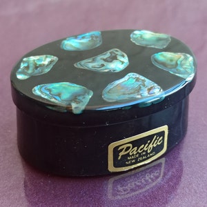 Puede incluir: Una caja de alhajas ovalada negra con una tapa decorada con piezas de concha de abulón azul y verde iridiscente. La caja tiene una etiqueta dorada que dice "Pacific Made in New Zealand".