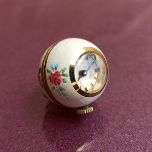 May include: A vintage, spherical watch with a cream-colored body and gold accents. The watch face is visible, and the body is decorated with a floral design in pink, blue, and green. The watch has a gold winding stem.