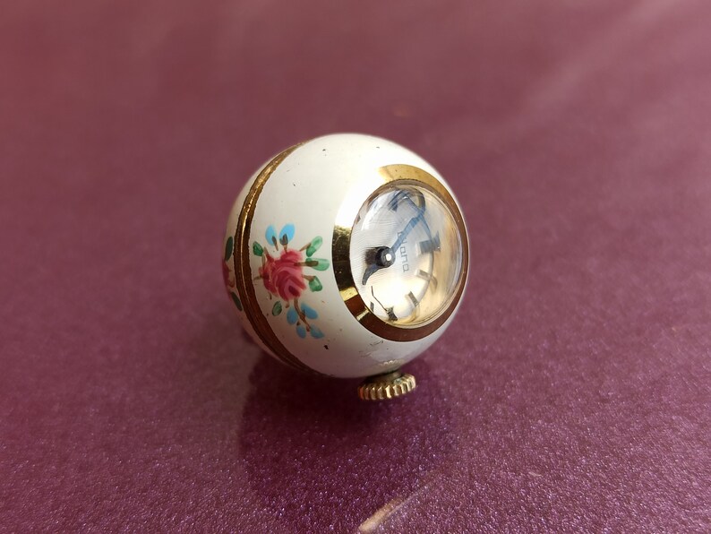May include: A vintage, spherical watch with a cream-colored body and gold accents. The watch face is visible, and the body is decorated with a floral design in pink, blue, and green. The watch has a gold winding stem.