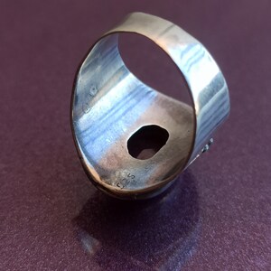 Pode incluir: Um anel de prata com uma pedra grande de forma ovalada engastada no centro. O anel &eacute; mostrado de lado, com a banda voltada para o espectador. A pedra est&aacute; faltando na engaste.