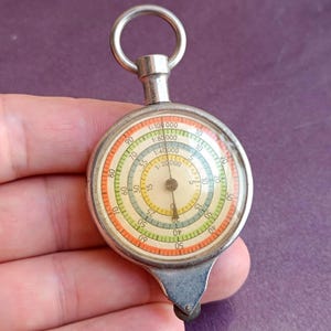 May include: A silver-toned map measuring tool featuring a circular face with multiple concentric scales in orange, green, and cream. The dial displays ratios including 1:20000, 1:40000, 1:80000, and 1:100000, with a single black needle and a top ring for hanging.