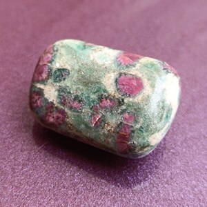 May include: A rounded, tumbled stone with a mix of green, white, and red hues. The stone's surface is smooth, with red inclusions creating a distinctive pattern. The stone is set against a purple background. The stone is likely a decorative item.