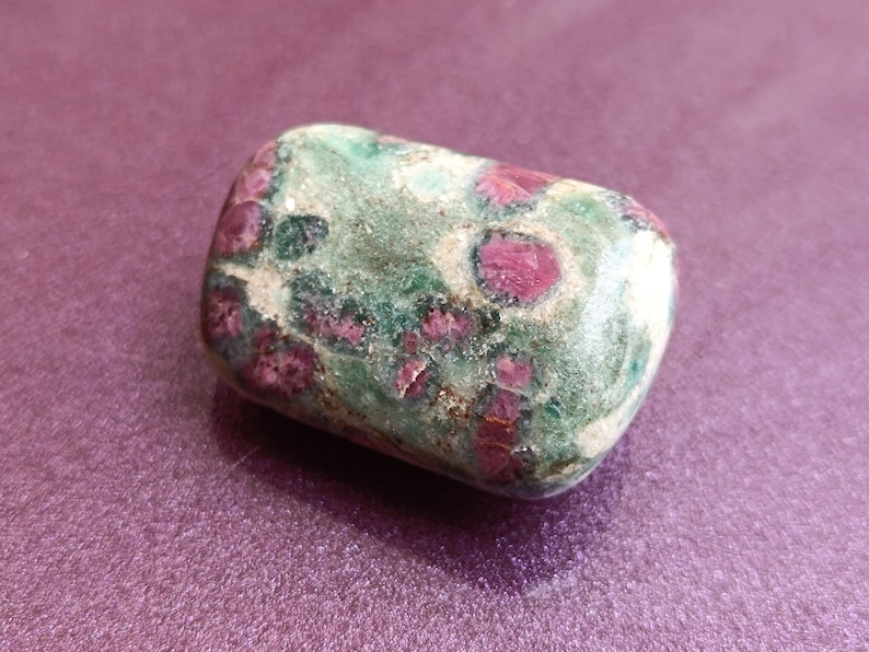 May include: A rounded, tumbled stone with a mix of green, white, and red hues. The stone's surface is smooth, with red inclusions creating a distinctive pattern. The stone is set against a purple background. The stone is likely a decorative item.