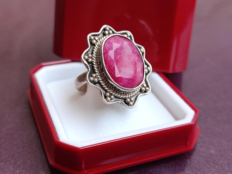 Op de afbeelding: Een zilveren ring met een grote, ovaal geslepen robijn. De robijn heeft een levendige roze kleur en is gezet in een decoratieve zilveren zetting met een gedetailleerde rand. De ring wordt gepresenteerd in een rode sieradendoos.