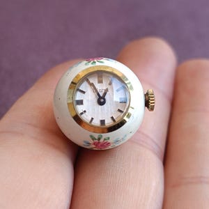 May include: A small, round vintage watch with a white face and gold-tone bezel. The watch has a floral design in pink and blue. The watch is held in a hand.
