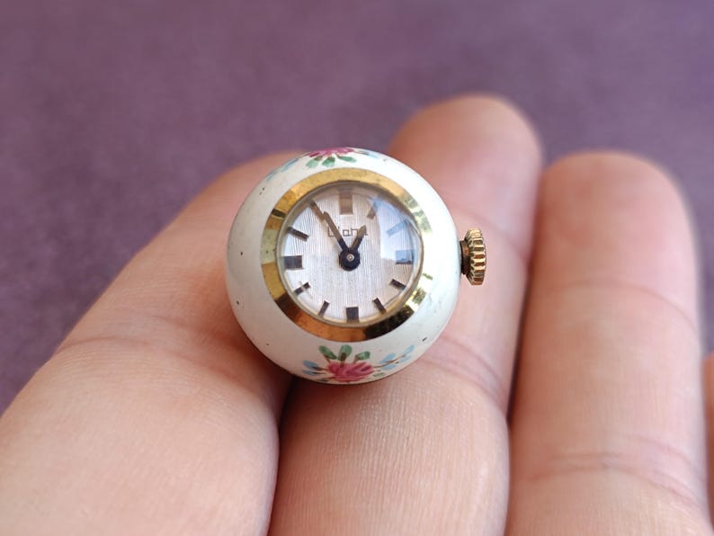 May include: A small, round vintage watch with a white face and gold-tone bezel. The watch has a floral design in pink and blue. The watch is held in a hand.