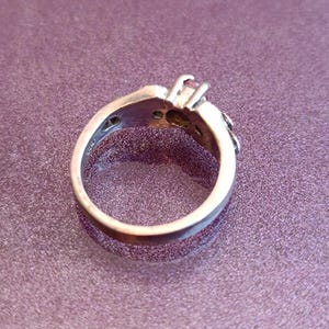 K&ouml;nnte beinhalten: Silberring mit einem zentralen rosa Edelstein und kleineren Akzentsteinen. Der Ring hat ein detailliertes Design um den Edelstein und entlang des Bandes. Der Ring ist vor einem violetten Hintergrund platziert.