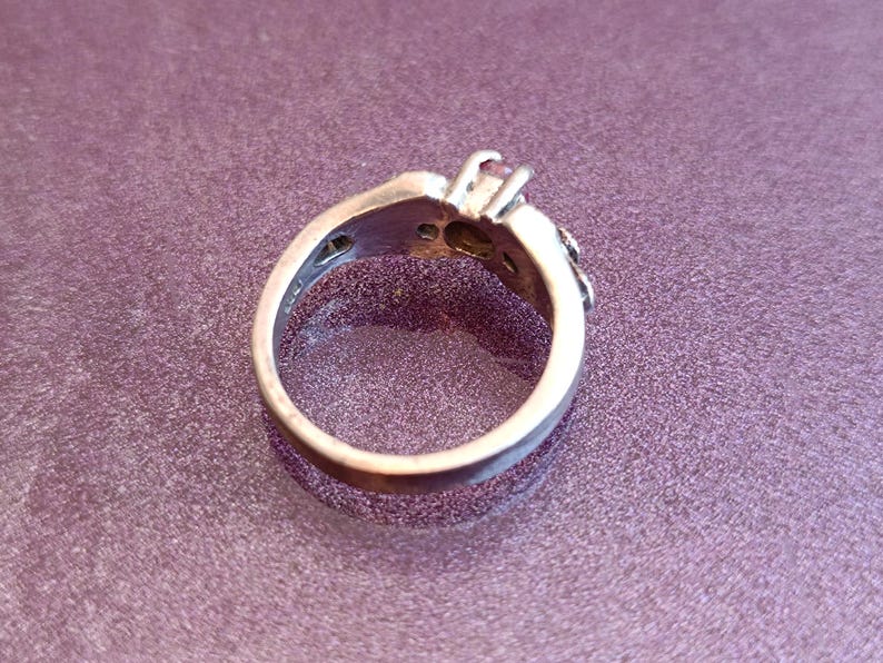 K&ouml;nnte beinhalten: Silberring mit einem zentralen rosa Edelstein und kleineren Akzentsteinen. Der Ring hat ein detailliertes Design um den Edelstein und entlang des Bandes. Der Ring ist vor einem violetten Hintergrund platziert.