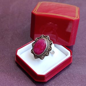 Op de afbeelding: Een zilveren ring met een grote, ovale, felroze edelsteen wordt getoond in een open rode sieradendoos. De ring heeft een decoratieve, kralenrand rond de steen. De doos is rood met een gouden rand.