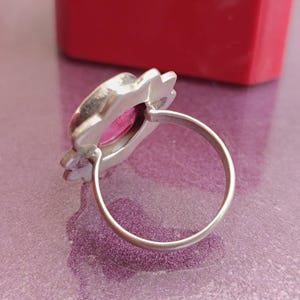 Op de afbeelding: Een zilveren ring met een roze edelsteen. De zetting van de ring heeft een bloemenontwerp rond de edelsteen. De ring wordt getoond op een paars oppervlak met een rood object op de achtergrond.