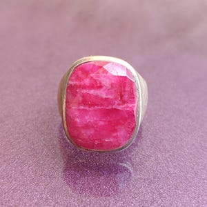 Op de afbeelding: Een zilveren ring met een grote, rechthoekige, gefacetteerde robijn edelsteen. De robijn heeft een levendige roze-rode kleur en de zilveren band heeft een eenvoudig, klassiek ontwerp. De ring staat tegen een paarse achtergrond.