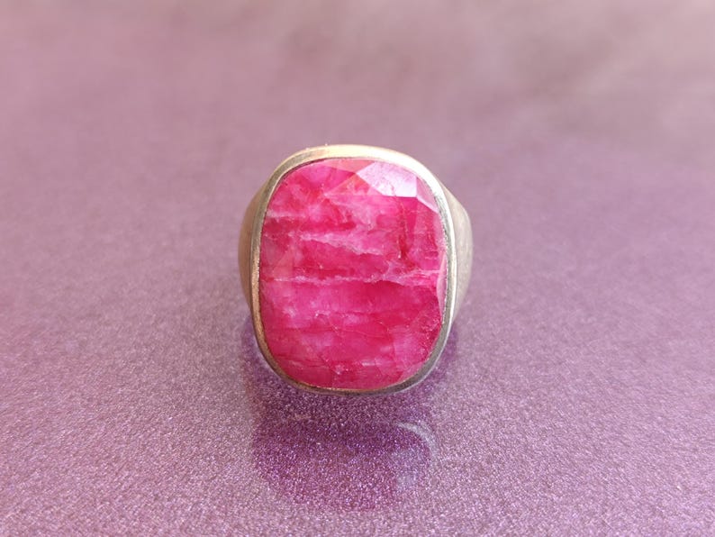 Op de afbeelding: Een zilveren ring met een grote, rechthoekige, gefacetteerde robijn edelsteen. De robijn heeft een levendige roze-rode kleur en de zilveren band heeft een eenvoudig, klassiek ontwerp. De ring staat tegen een paarse achtergrond.