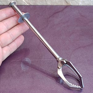 Può includere: Uno strumento di presa in metallo color argento dotato di un'asta lunga e sottile, un'impugnatura a stantuffo a un'estremità e quattro artigli di presa retrattili e seghettati all'altra, progettato per recuperare piccoli oggetti in spazi stretti.