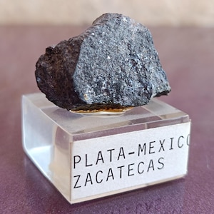 May include: A dark gray mineral specimen, possibly a rock or ore, sits atop a clear acrylic display stand. The stand features a label with the text "PLATA-MEXICO ZACATECAS". The mineral has a rough, textured surface.