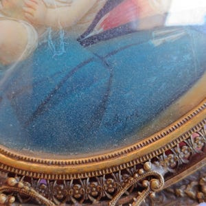 K&ouml;nnte beinhalten: Antikes ovales Portr&auml;t in einem dekorativen goldfarbenen Rahmen. Das Kunstwerk zeigt eine religi&ouml;se Szene mit Figuren in Blau, Rot und Wei&szlig;. Der Rahmen hat aufwendige Blumen- und Schn&ouml;rkeldetails, die seinen Vintage-Charme unterstreichen.