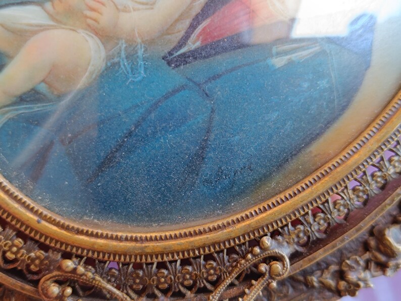 K&ouml;nnte beinhalten: Antikes ovales Portr&auml;t in einem dekorativen goldfarbenen Rahmen. Das Kunstwerk zeigt eine religi&ouml;se Szene mit Figuren in Blau, Rot und Wei&szlig;. Der Rahmen hat aufwendige Blumen- und Schn&ouml;rkeldetails, die seinen Vintage-Charme unterstreichen.