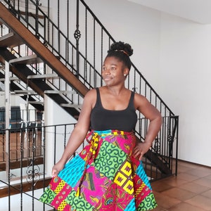 Könnte beinhalten: Eine Frau trägt ein schwarzes Tanktop und einen bunten Wickelrock mit geometrischem Muster. Der Rock ist aus einem leuchtenden Stoff in den Farben Pink, Grün, Gelb und Blau. Sie steht vor einer Treppe mit einem schwarzen Geländer.