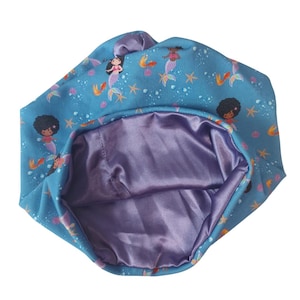 Könnte beinhalten: Eine blaue, satin-gefütterte Schlafhaube mit einem Meerjungfrauen-Druck, der schwarze Mädchen mit braunen Haaren zeigt. Die Haube hat einen gerafften Oberteil und ist so konzipiert, dass sie das Haar während des Schlafs schützt.