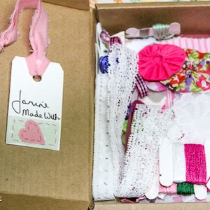 May include: A box filled with pink and white lace, fabric scraps, and embroidery floss. The box has a tag attached that says "Janine Made With" and a pink heart.