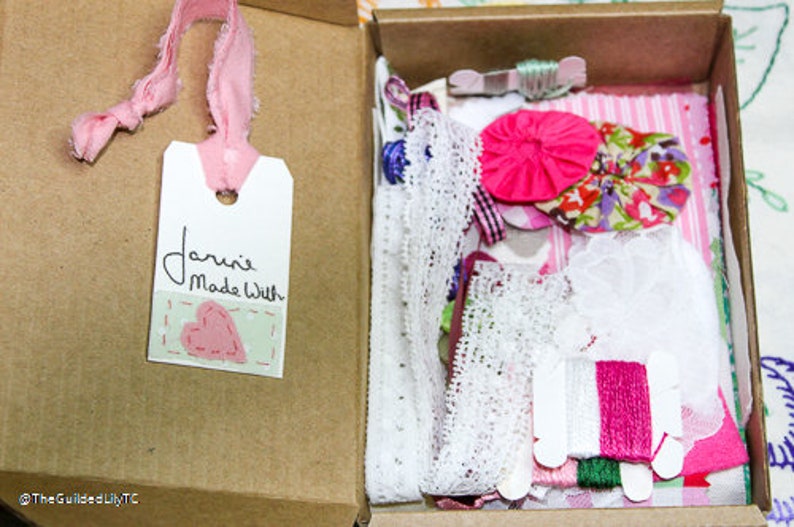 May include: A box filled with pink and white lace, fabric scraps, and embroidery floss. The box has a tag attached that says "Janine Made With" and a pink heart.