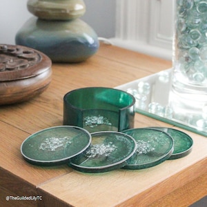 Op de afbeelding: Een set van vier groene hars onderzetters met zilveren accenten en een bijpassende houder. De onderzetters zijn rond met een zilveren rand. De set wordt gepresenteerd op een houten oppervlak.