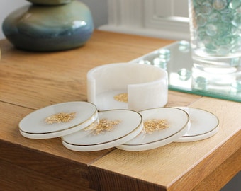 May include: A set of white resin coasters with gold flecks and gold trim, along with a matching holder. The coasters are round and sit on a wooden table. The set is designed for home decor.