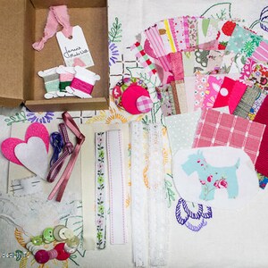 May include: A craft kit with a variety of fabrics, trims, and embellishments in pink, white, and green. The kit includes two felt hearts, a lace trim, a ribbon, and a small piece of fabric with a dog design. The kit also includes a tag that says "Jane Madish".