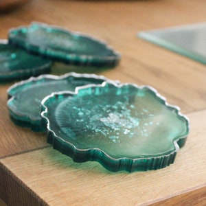 May include: Four teal green coasters with a silver rim. The coasters are stacked on top of each other. The coasters have a unique, organic shape.