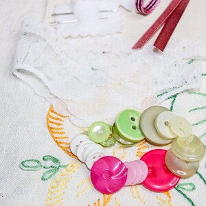 May include: A collection of assorted buttons in various colors and sizes, including green, white, and brown. The buttons are arranged on a white fabric background with lace and embroidery details.