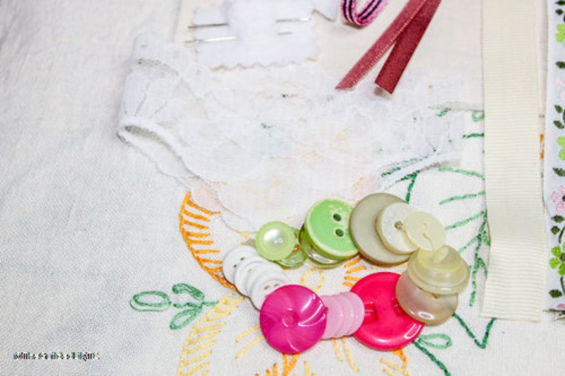 May include: A collection of assorted buttons in various colors and sizes, including green, white, and brown. The buttons are arranged on a white fabric background with lace and embroidery details.