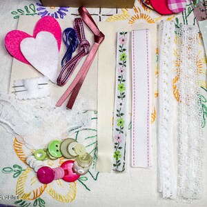 May include: A collection of craft supplies including white lace, pink ribbon, pink felt hearts, and green and white buttons.