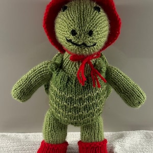 May include: A green knitted turtle doll wearing a red knitted hat, a red bow, and red booties.