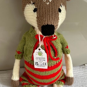 Pode incluir: Um brinquedo de rena de malha que veste um vestido listrado verde e vermelho com um suéter verde e uma fita rosa. O brinquedo tem chifres marrons, pernas marrons e um rosto branco com olhos pretos.