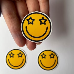Puede incluir: Tres pegatinas amarillas con forma de cara sonriente con contornos negros y ojos en forma de estrella. Las pegatinas están sobre una superficie blanca.