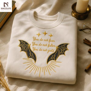 May include: Cream-colored sweatshirt with embroidered design. Features a bat wing motif in black and gold, with the text "You do not fear, You do not falter, You do not yield." Three gold stars are above the text. A sunburst design is below the text.