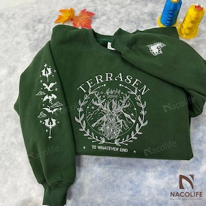 Peut inclure: Un sweat-shirt vert foncé avec un motif graphique blanc représentant un cerf avec des bois et une couronne. Le texte "TERRASEN TO WHATEVER END" est imprimé sous le cerf. Le sweat-shirt a un motif de dragon blanc sur la manche.