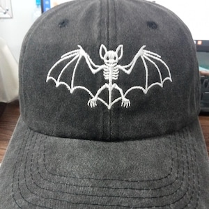May include: A charcoal gray baseball cap featuring an embroidered white graphic of a bat skeleton with outstretched wings centered on the front panel. The cap has a curved brim with visible stitching and a button at the top of the crown.