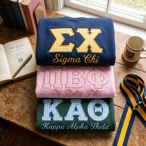 May include: Three folded crewneck sweatshirts stacked vertically. The top is navy blue with yellow gingham Sigma Chi letters, the middle is light pink with pink gingham Pi Beta Phi letters, and the bottom is dark green with blue gingham Kappa Alpha Theta letters.